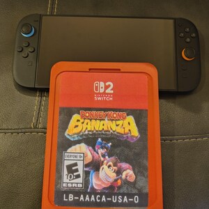 Puede incluir: Una consola Nintendo Switch negra con mandos azules y naranjas se muestra encima de un cartucho de juego rojo. El cartucho presenta el título del juego "Donkey Kong Bananza" y la clasificación ESRB "Everyone 10+".