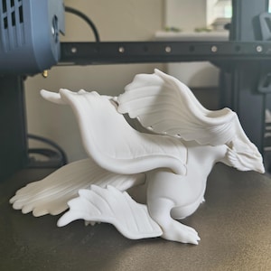 May include: A white, 3D-printed dragon figurine with detailed wings and a stylized body. The dragon is posed in a crouching position, showcasing its wingspan. The figurine is likely a decorative item or a collectible.