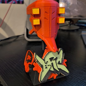 以下が含まれることがあります： 黒色の台座に、赤と黄色の3Dプリントされたロボットの頭部の置物。台座には緑と赤の落書きのデザインが施されている。ロボットの頭部には、側面に黄色の正方形が4つある。