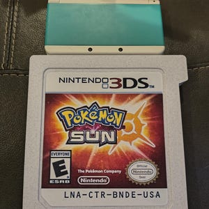 May include: A teal Nintendo 3DS console rests above a game cartridge for Pokémon Sun. The game cartridge features the Pokémon Sun logo with a sunburst design and the Nintendo and ESRB logos. The cartridge has the text "LNA-CTR-BNDE-USA".