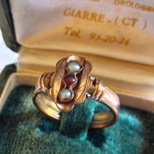 Può includere: Un anello d'oro dal design ornato, con tre pietre incastonate: due perle e una gemma rossa centrale. L'anello è esposto in un portagioie foderato in velluto verde, con l'interno della scatola visibile. L'anello presenta dettagli intricati.