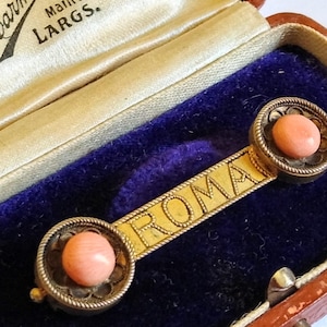 Peut inclure: Broche vintage dorée avec le mot "ROMA" gravé sur une barre. La broche comporte deux montures circulaires, chacune contenant une pierre rose corail. La broche est présentée dans un écrin à bijoux marron avec un intérieur en velours bleu.