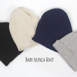 May include: Four knit beanies in black, cream, navy blue, and light gray. Each hat features a ribbed texture and a folded cuff. The text "BABY ALPACA KNIT" is printed in black in the center of the image, positioned below the four beanies.