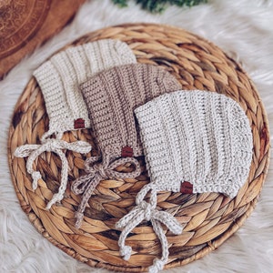 May include: Three crocheted baby bonnets in beige, brown, and white with matching ties. The bonnets are arranged on a woven straw mat.