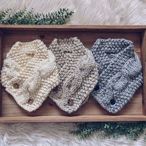 May include: Three knitted neck warmers in a wooden tray. The neck warmers are in cream, beige, and gray with a cable knit pattern and wooden buttons.