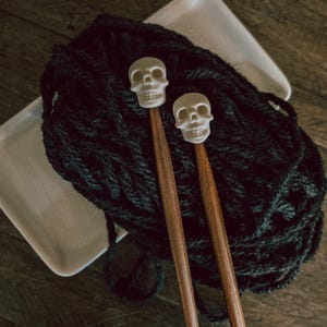 May include: Two wooden knitting needles with white skull-shaped tips. The needles are resting on a ball of black yarn.