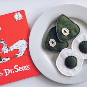 May include: A white plate with three felt food items: two green eggs and one green ham. The plate is next to a copy of the book "Green Eggs and Ham" by Dr. Seuss.