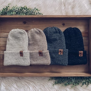 May include: Four knitted beanies in a wooden tray. The beanies are in beige, light brown, dark gray, and black. Each beanie has a small leather patch with a bear design.
