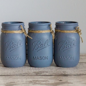 May include: Three blue glass mason jars with a brown twine bow around the top of each jar. The jars are labeled "Ball Mason" and are sitting on a wooden surface.