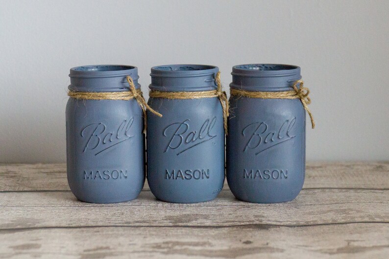May include: Three blue glass mason jars with a brown twine bow around the top of each jar. The jars are labeled "Ball Mason" and are sitting on a wooden surface.