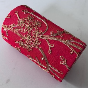 Puede incluir: Un objeto cilíndrico de tela roja con bordados dorados. El bordado representa un diseño floral y de ramas. El artículo parece ser un contenedor o funda decorativa, posiblemente para un objeto pequeño.