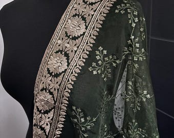 Puede incluir: Chale bordado verde oscuro con diseño floral. El chal tiene un borde ancho con intrincados patrones florales dorados. La tela es transparente y tiene una apariencia delicada y brillante. El chal está drapeado sobre un maniquí negro.
