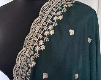 Puede incluir: Un chal verde oscuro con intrincados bordados dorados en los bordes y dispersos por la tela. El bordado presenta patrones florales y geométricos, creando un aspecto lujoso y elegante. El chal está drapeado sobre un maniquí negro.