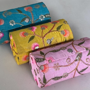 May include: Three rectangular fabric cases in teal, yellow, and pink. Each case has floral embroidery with pink, orange, and green accents, silver sequins, and a silver button closure. The cases are arranged at a slight angle, showing their details.
