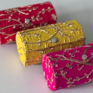 Puede incluir: Tres cajas rectangulares de tela en rojo, amarillo y fucsia. Cada caja está adornada con patrones florales bordados en color dorado y un cierre de botón plateado. Las cajas están dispuestas en fila. Ideales para joyas.