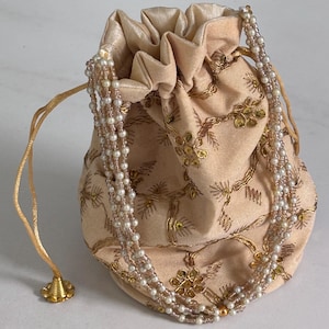 以下が含まれることがあります： 金色の刺繍とビーズのネックレスが付いた、桃色の巾着袋。巾着袋はギャザーの入った上部と、紐の付け根にある金のタッセルが特徴です。ネックレスは白とピンクのビーズでできています。