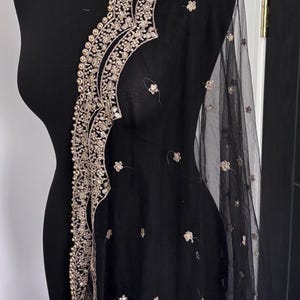 May include: Black sheer shawl with intricate gold embroidery along the edges. The shawl is draped over a black mannequin, showcasing the delicate floral and beaded details. The design features a scalloped edge and scattered floral accents.