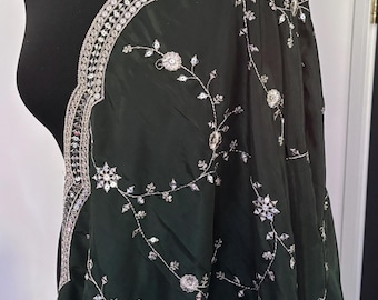 Puede incluir: Un chal de color verde oscuro con un borde bordado en plata y motivos florales plateados dispersos. La tela tiene un brillo sutil y el bordado incluye pequeñas lentejuelas reflectantes que captan la luz en toda la superficie.