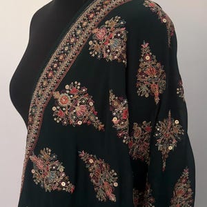 May include: A dark green shawl with detailed floral embroidery in red, pink, and gold. The shawl is draped over a black mannequin, highlighting the intricate design and texture. The embroidery includes sequins.