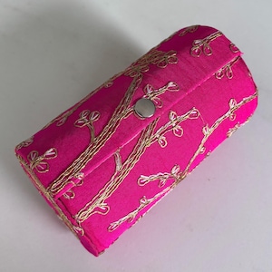 Puede incluir: Un estuche cilíndrico de color rosa brillante con cierre de solapa. El estuche está adornado con patrones florales bordados en color dorado. Un broche plateado asegura la solapa. El estuche parece estar hecho de tela.