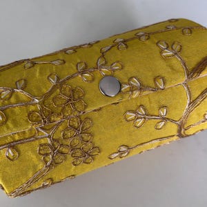 Puede incluir: Un bolso de mano de tela amarilla con un diseño de bordado floral en marrón y blanco. El bolso de mano tiene forma redondeada y un cierre de botón a presión plateado. El bordado presenta un patrón de ramas de árbol.