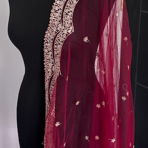 Puede incluir: Un dupatta burdeos, drapeado sobre un maniquí negro. El borde presenta intrincados bordados dorados y pequeños motivos florales dorados dispersos. La tela transparente añade un toque de elegancia a esta prenda tradicional.