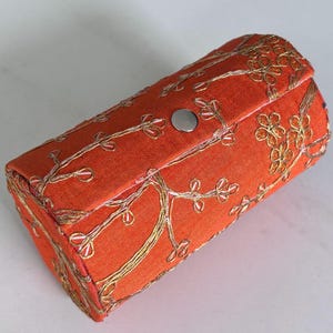 Puede incluir: Caja cilíndrica de tela naranja con bordado floral dorado y cierre de botón a presión plateado. El diseño bordado presenta ramas y flores, creando un patrón elegante y ornamentado. La caja mide aproximadamente 15 cm de largo.