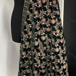 Puede incluir: Un chal de terciopelo verde oscuro con un intrincado diseño floral y un borde decorativo. El chal está drapeado sobre un maniquí negro, mostrando su patrón y textura. El diseño floral incluye detalles dorados, rosas y blancos.