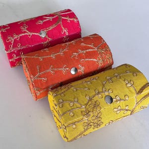 Puede incluir: Tres cajas rectangulares de tela en rosa, naranja y amarillo, cada una con la parte superior curvada. Cuentan con diseños florales bordados en dorado y un cierre de botón plateado. Estas cajas son probablemente para guardar joyas u objetos pequeños.