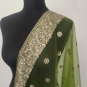 May include: A green sheer scarf with gold and silver sequins and mirror work. The scarf has a floral design and is trimmed with a border of sequins.