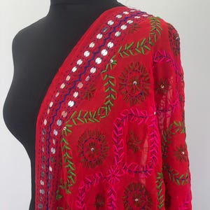 May include: A red sheer shawl with intricate embroidery and silver mirror work. The shawl features a floral design with green, pink, and blue thread.