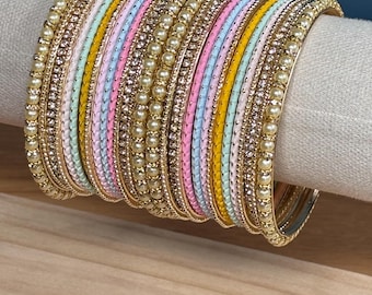 Może przedstawiać: Zestaw ułożonych w stos bransoletek typu bangle, zawierający różnorodne emaliowane paski w pastelowych odcieniach różu, żółci i błękitu, przeplatane złotymi metalowymi paskami ozdobionymi małymi białymi sztucznymi perłami i błyszczącymi cyrkoniami.