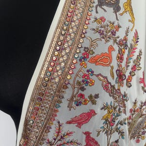 Peut inclure: Écharpe blanche avec un fond bleu clair et des broderies complexes. La bordure est ornée de paillettes dorées et de broderies colorées d'oiseaux, de fleurs et d'animaux. Le design est détaillé et orné.