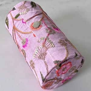 May include: A cylindrical, lavender-colored case with intricate floral embroidery in shades of pink, gold, and green. The design includes sequins and a silver button closure. The case appears to be made of fabric.