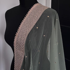 May include: A sheer, pale green scarf with a decorative border. The border is a light pink with a lattice design and small, silver-colored beads. Scattered gold-colored sequins add sparkle to the fabric.