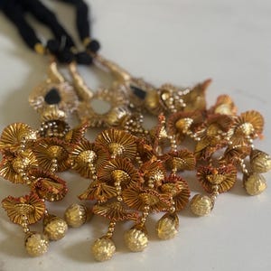 May include: Close-up of ornate gold-colored tassels with intricate floral designs and dangling spherical beads. The tassels are attached to black cords. The details include a mix of textures and reflective surfaces, suggesting a handcrafted item.