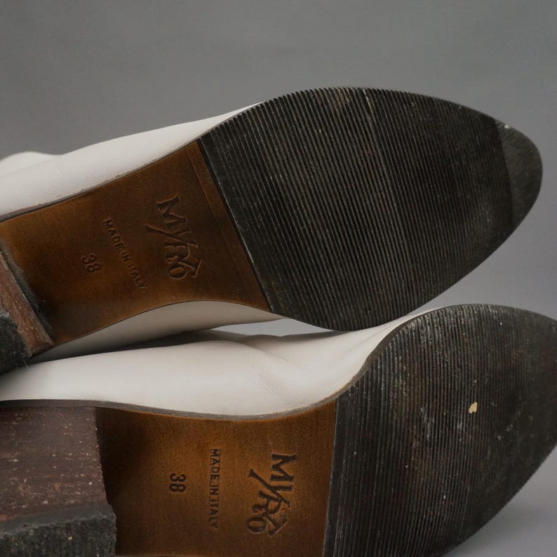 Puede incluir: Primer plano de las suelas de dos botas de cuero blanco. Las suelas son de color marr&oacute;n oscuro con un patr&oacute;n texturizado y acanalado. Los tacones tambi&eacute;n son de color marr&oacute;n oscuro. Las botas est&aacute;n marcadas "Made in Italy" y talla 38.