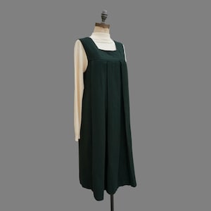 May include: A dark green jumper dress with a square neckline and pleated front, worn over a cream-colored long-sleeved top. The dress is displayed on a mannequin against a neutral gray background. The overall style is vintage.