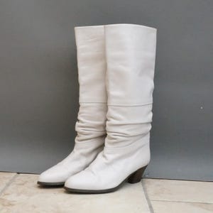 Puede incluir: Un par de botas altas de color blanco roto. Las botas tienen un dise&ntilde;o holgado con una textura ligeramente arrugada. Cuentan con una puntera redondeada y un tac&oacute;n de bloque marr&oacute;n oscuro.