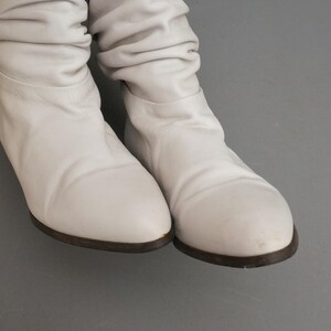 Puede incluir: Un par de botas de cuero blanco con punta redondeada y suela baja marr&oacute;n oscuro. Las botas tienen un dise&ntilde;o holgado, con el cuero fruncido alrededor de los tobillos. El fondo es gris liso.