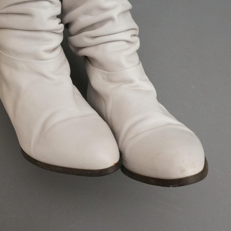 Puede incluir: Un par de botas de cuero blanco con punta redondeada y suela baja marr&oacute;n oscuro. Las botas tienen un dise&ntilde;o holgado, con el cuero fruncido alrededor de los tobillos. El fondo es gris liso.
