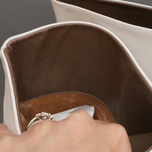 Puede incluir: Primer plano de un bolso de cuero blanco con interior marr&oacute;n. Una mano se introduce en el bolso, sosteniendo un anillo de oro con tres piedras. El anillo est&aacute; sobre papel blanco.