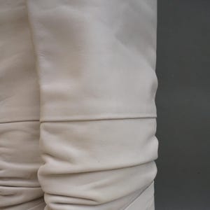 Puede incluir: Primer plano de botas de cuero blanco roto. El cuero es suave y fruncido, con un detalle de costura horizontal. Las botas tienen un color neutro y una textura suave, lo que sugiere un material de alta calidad.
