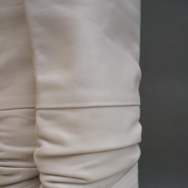 Puede incluir: Primer plano de botas de cuero blanco roto. El cuero es suave y fruncido, con un detalle de costura horizontal. Las botas tienen un color neutro y una textura suave, lo que sugiere un material de alta calidad.