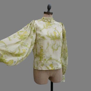 Może przedstawiać: Bluzka w stylu vintage z wysokim kołnierzem z falbankami i długimi, bufiastymi rękawami. Tkanina ma kremowe tło z zielono-żółtym wzorem kwiatowym. Bluzka jest prezentowana na manekinie.