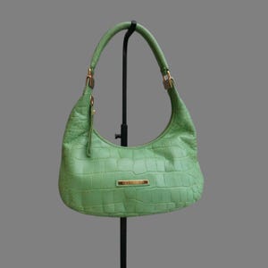 May include: A light green, crocodile-textured handbag with a curved shape and a single handle. The bag has gold-colored hardware and a small rectangular plaque on the front. The bag is displayed on a black stand.