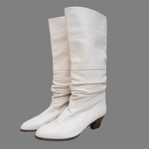Puede incluir: Un par de botas blancas de cuero hasta la rodilla con un dise&ntilde;o ligeramente arrugado. Las botas tienen un tac&oacute;n bajo de color marr&oacute;n oscuro y una puntera redondeada. Las botas miden aproximadamente 38 cm de alto.