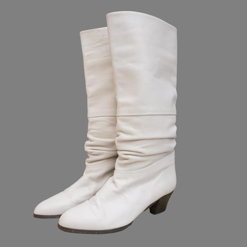 Puede incluir: Un par de botas blancas de cuero hasta la rodilla con un dise&ntilde;o ligeramente arrugado. Las botas tienen un tac&oacute;n bajo de color marr&oacute;n oscuro y una puntera redondeada. Las botas miden aproximadamente 38 cm de alto.