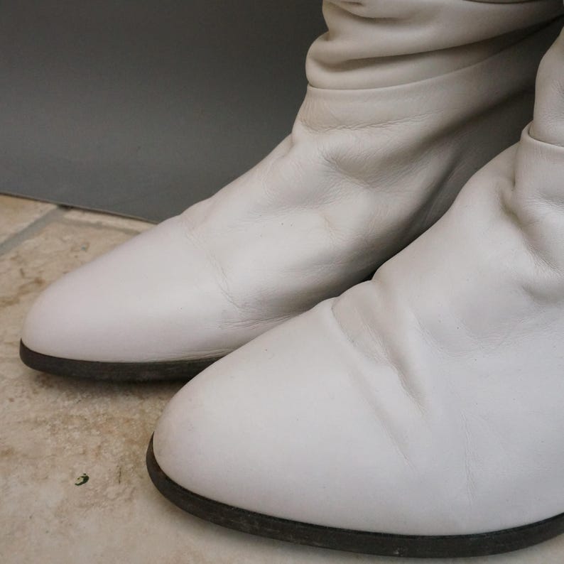 Puede incluir: Un par de botas de cuero blanco con punta redondeada y suela marr&oacute;n oscuro. Las botas tienen una textura ligeramente arrugada y una altura a media pantorrilla. Las botas est&aacute;n sobre un fondo neutro.
