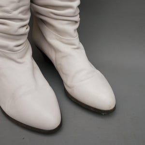 Puede incluir: Un par de botas de cuero blanco roto con un dise&ntilde;o arrugado en la parte superior. Las botas tienen un tac&oacute;n bajo y una punta redondeada. Las suelas son de color marr&oacute;n oscuro. Las botas se muestran sobre un fondo gris.
